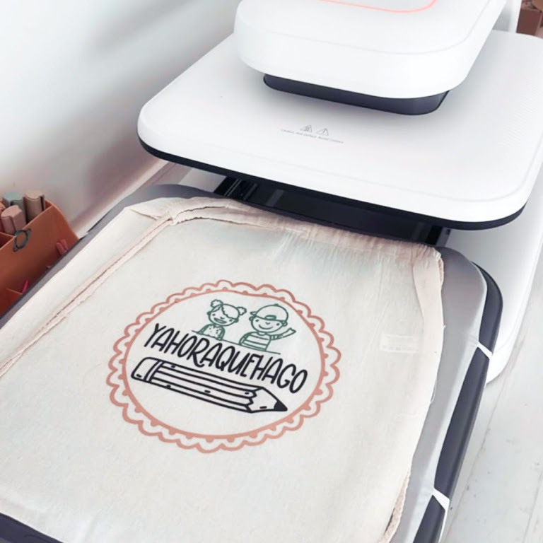 LOKLiK ImPress™ Auto 2-Smart in Daisy White. On the ironing board is a drawstring canvas bag with a heat transfer vinyl of a circle. Withint the circle is a line illustration of two children and apencil and the word 'yahoraquehago'.