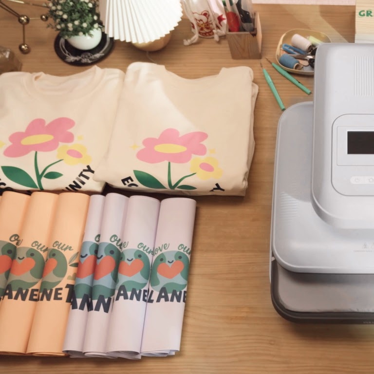 A LOKLiK ImPress™ Auto 2-Smart on a wooden table. On the left is a stack of folded T-shirts with designs in heat transfer vinyl
