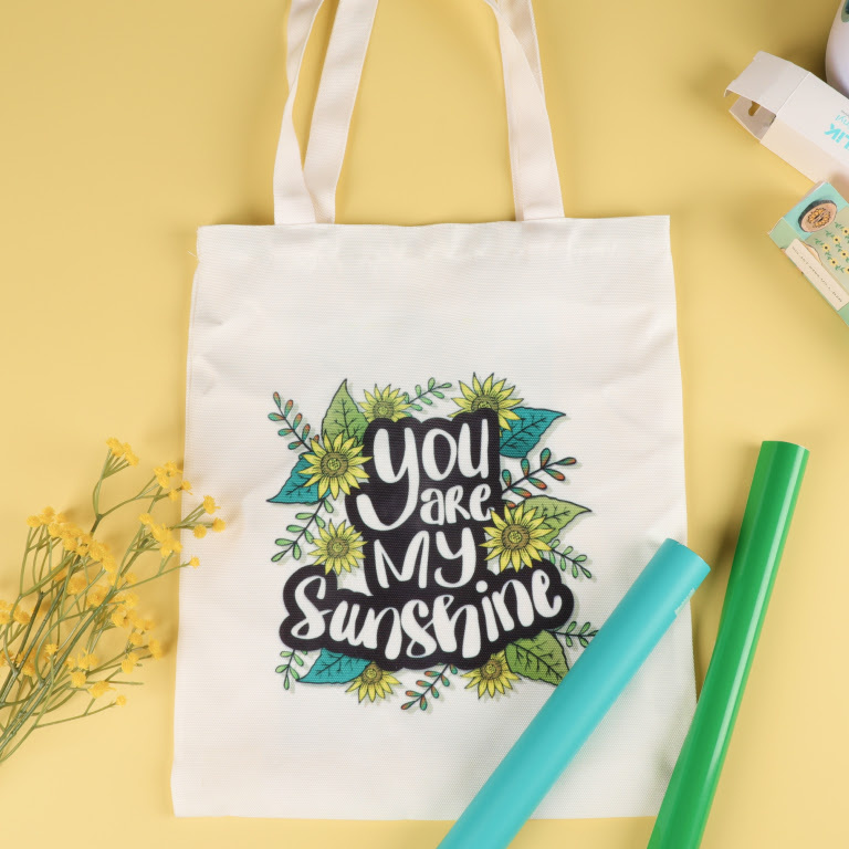 Top down view of a white canvas tote bag against a light pink background. The tote bag has a heat transfer design of flowers and the text 'You are my sunshine' 