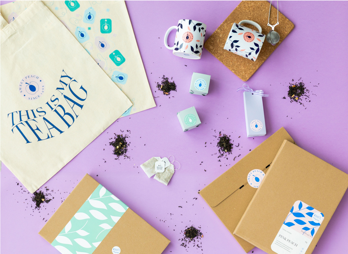 Ansicht von oben auf eine individuell gestaltete Kartonverpackung, individuell gestaltete kleine Schachteln und Tassen mit einem Sublimationsdesign und zwei Tragetaschen mit einem Design aus Teebeuteln und dem Text "Dies ist mein Teebeutel" vor einem hellvioletten Hintergrund.
