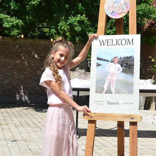 Young girl proudly standing next to an easel with a big piece of cardboard. It says welcome and has a picture of the girl.