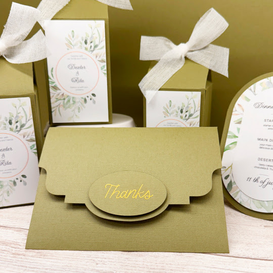 Envelope for a wedding invitation in olive coloured paper and with an embellishment in gold foil saying 'Thanks'