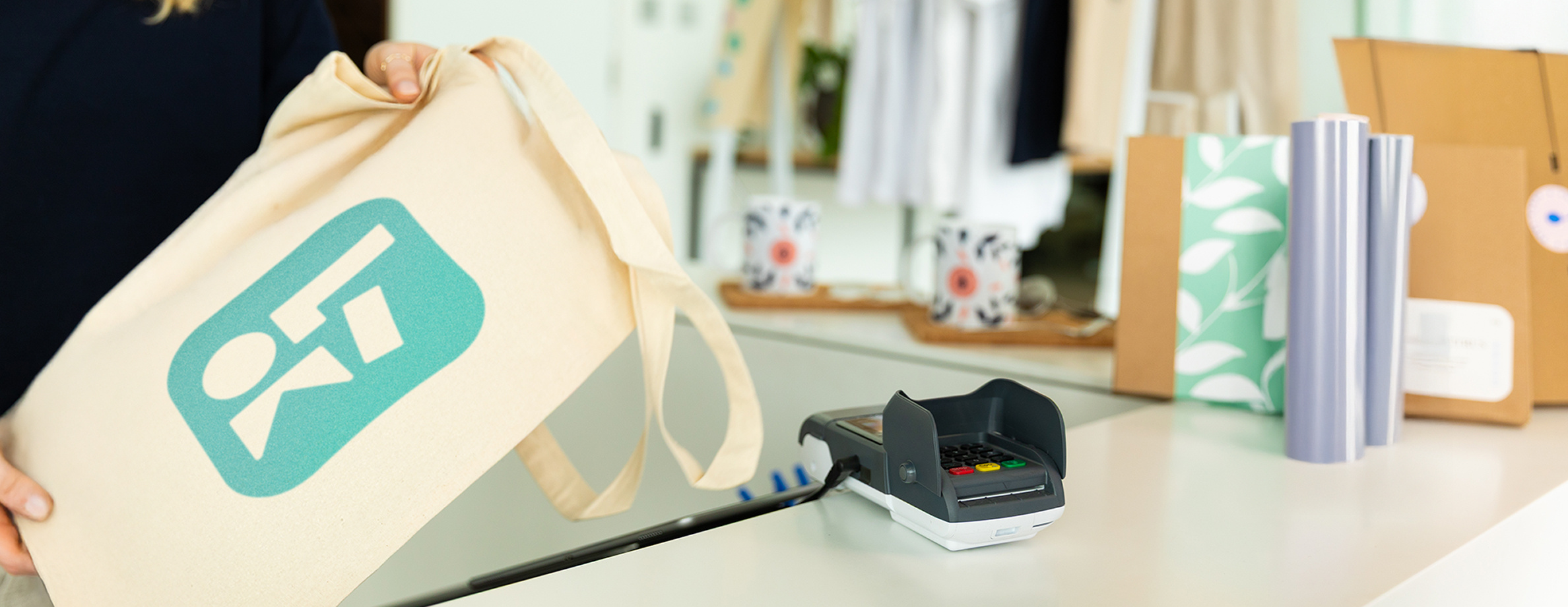 Betaalterminal op een witte toonbank. Links houdt een vrouw een canvas draagtas met het LOKLiK logo vast. Rechts staan een paar rollen vinyl rechtop en kartonnen verpakkingen.