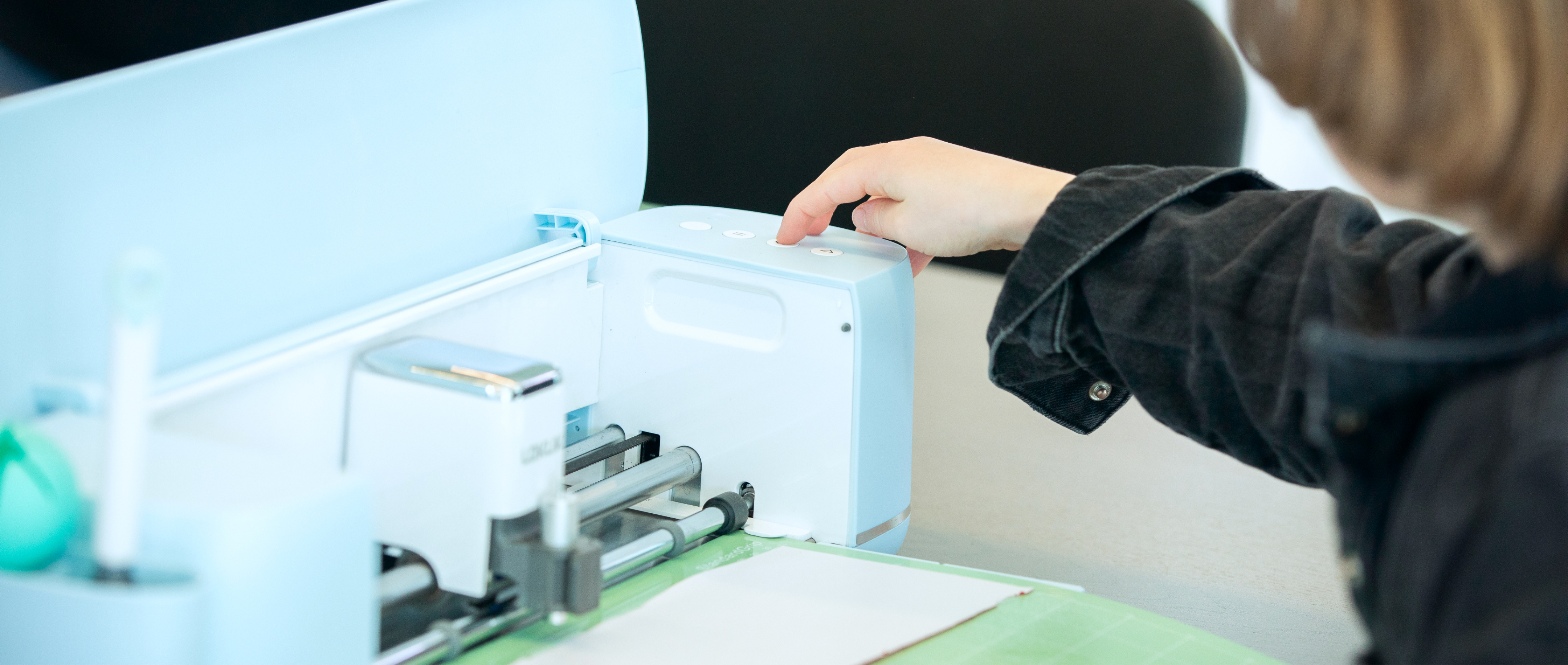 Enfant vêtu d'une veste en jean noire appuyant sur le bouton de démarrage d'un LOKLiK iCraft™ en Bubble Blue. Un tapis de découpe vert avec du vinyle a été chargé.