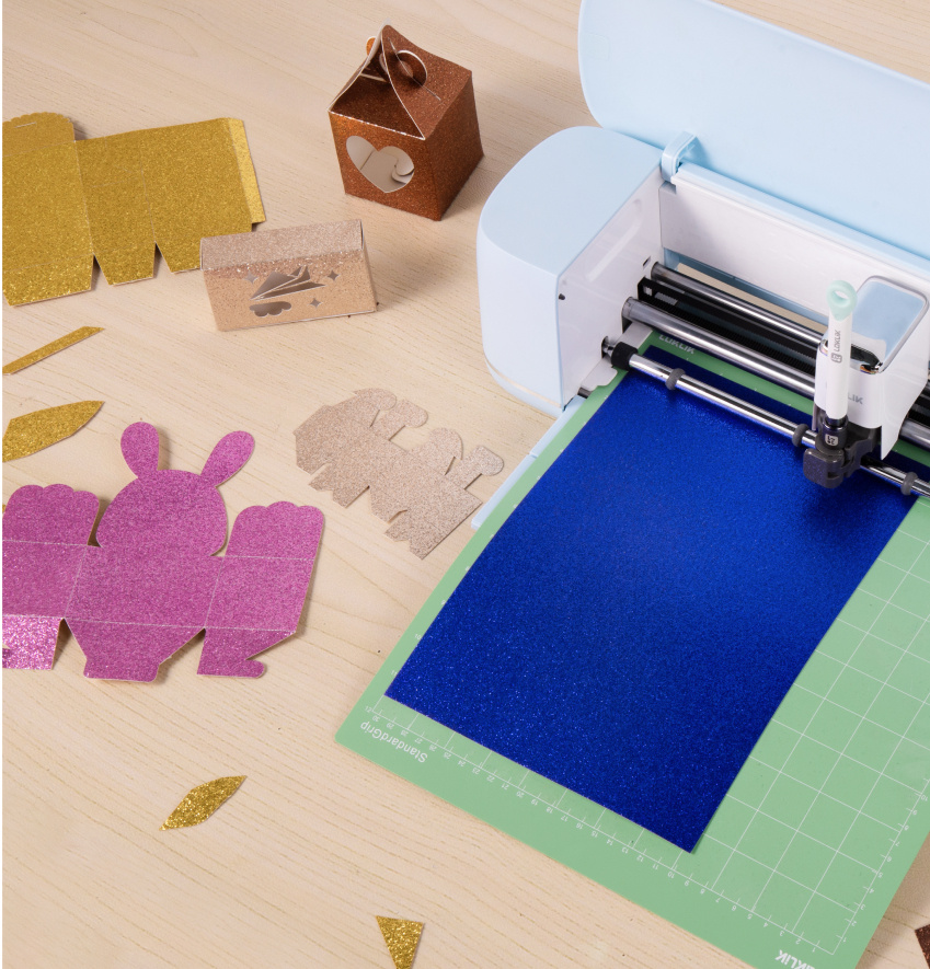 LOKLiK iCraft™ en Bubble Blue sur une table en bois. Un tapis de découpe vert avec du papier cartonné bleu est chargé dans la machine de découpe. À côté se trouvent plusieurs petites boîtes plates qui ont été découpées.