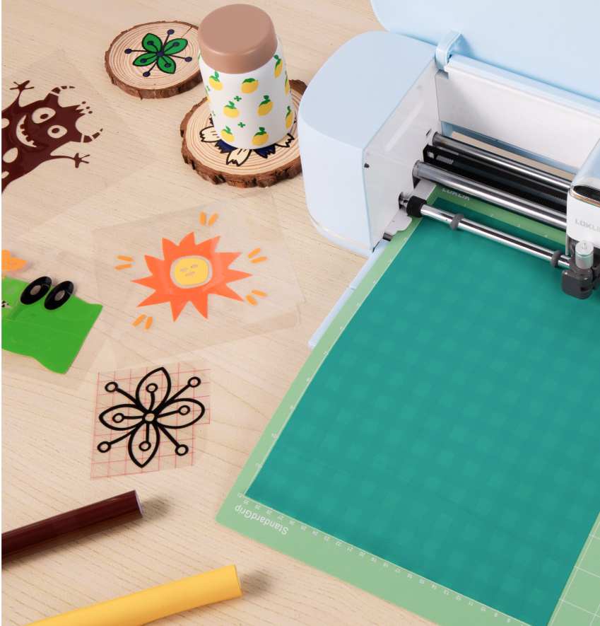 LOKLiK iCraft™ en Bubble Blue sur une table en bois. Un tapis de découpe vert avec du vinyle adhésif permanent a été chargé dans la machine de découpe. À côté se trouvent plusieurs motifs en vinyle adhésif permanent déjà découpés.