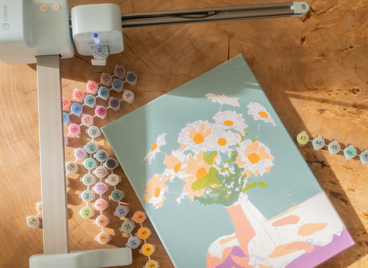 Bovenaanzicht van een gedeeltelijk geschilderd paint-by numbers kunstwerk op een doek tegen een houten ondergrond. Het stelt een vaas met bloemen voor. Rondom het doek staan kleine genummerde potjes verf. Een LOKLiK iPaint™ omlijst het tafereel.