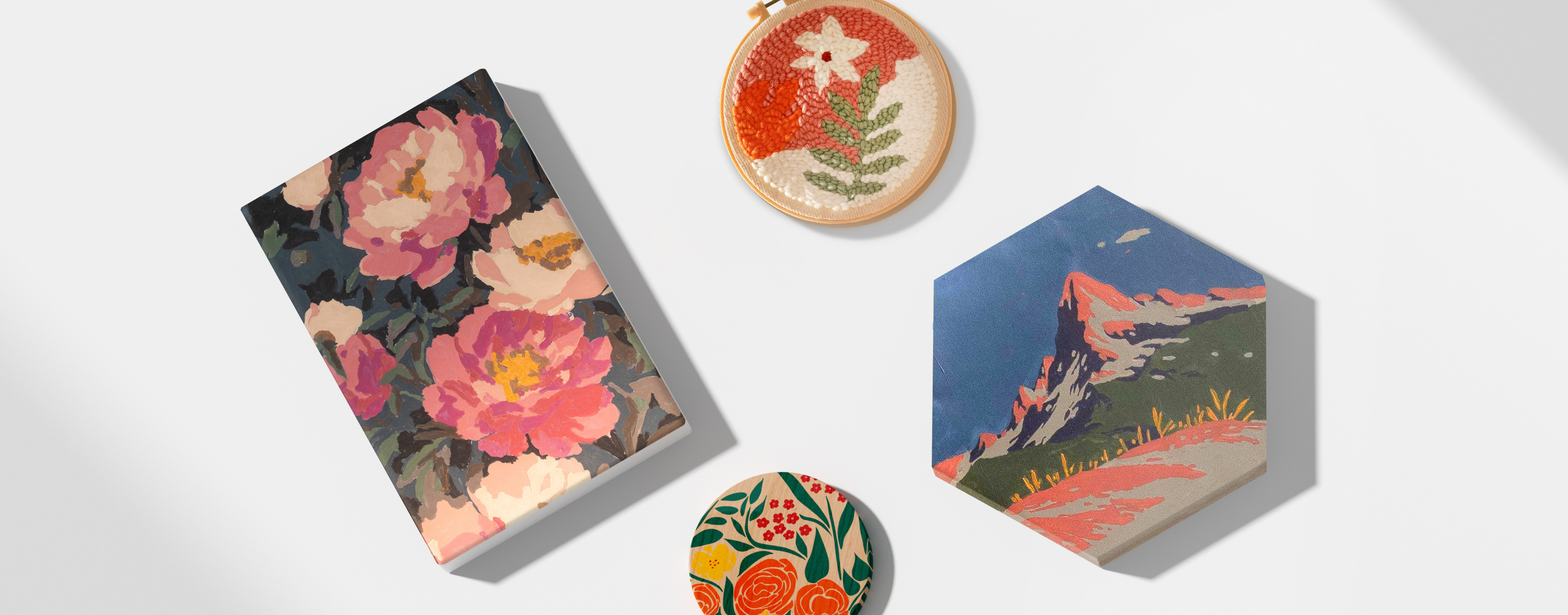 Bovenaanzicht van een wit vlak met een afgewerkt paint-by-numbers canvas van bloemen, een zeshoekig canvas met een paint-by-numbers schildering van een berg, een borduurring met een getuft ontwerp van een witte bloem en groen blad tegen een roze en witte achtergrond en een kleine houten onderzetter met een geschilderd ontwerp van bloemen en bladeren.