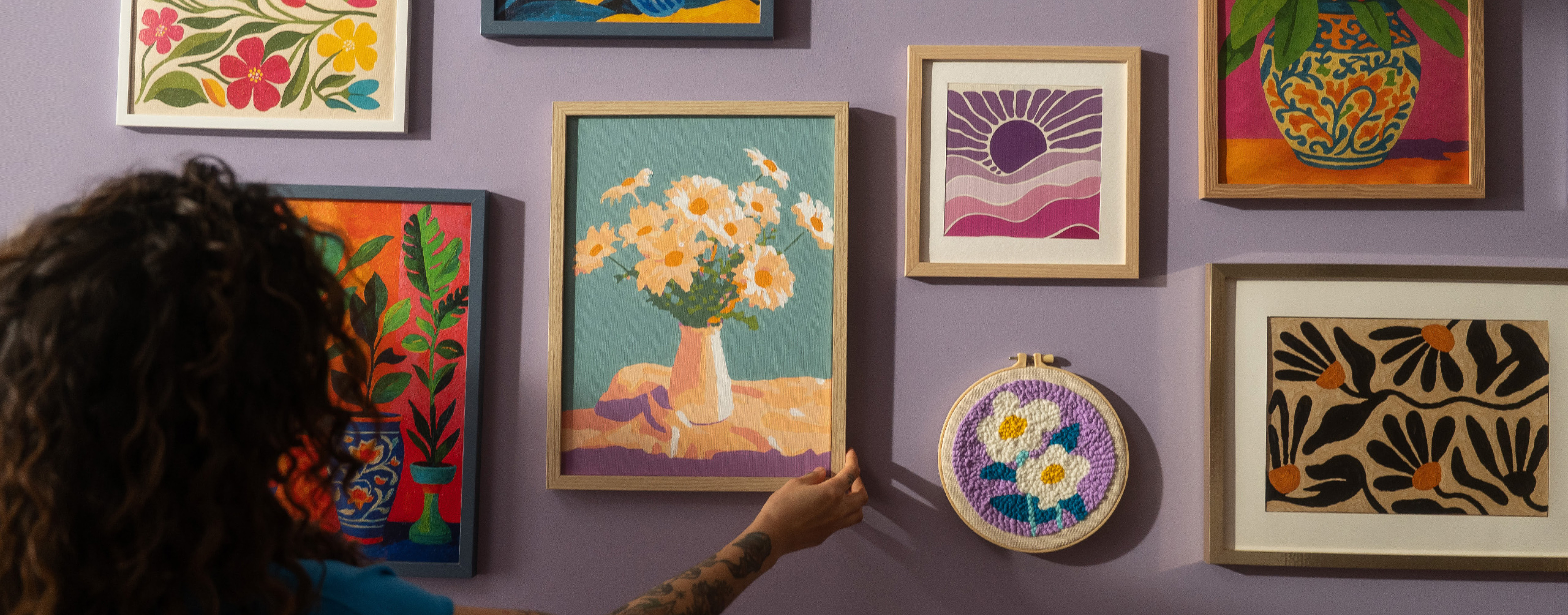 Vrouw verstelt een lijst met een paint-by-numberschilderij van een vaas met witte bloemen. De paarse muur is versierd met verschillende andere paint-by-numbers kunstwerken en een borduurring met een getuft motief van twee witte bloemen tegen een paarse achtergrond.