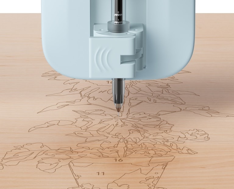LOKLiK gelpen in de LOKLiK iPaint™ penhouder. De pen tekent een paint-by-numbers van een zonnebloem in een pot op een houten oppervlak.