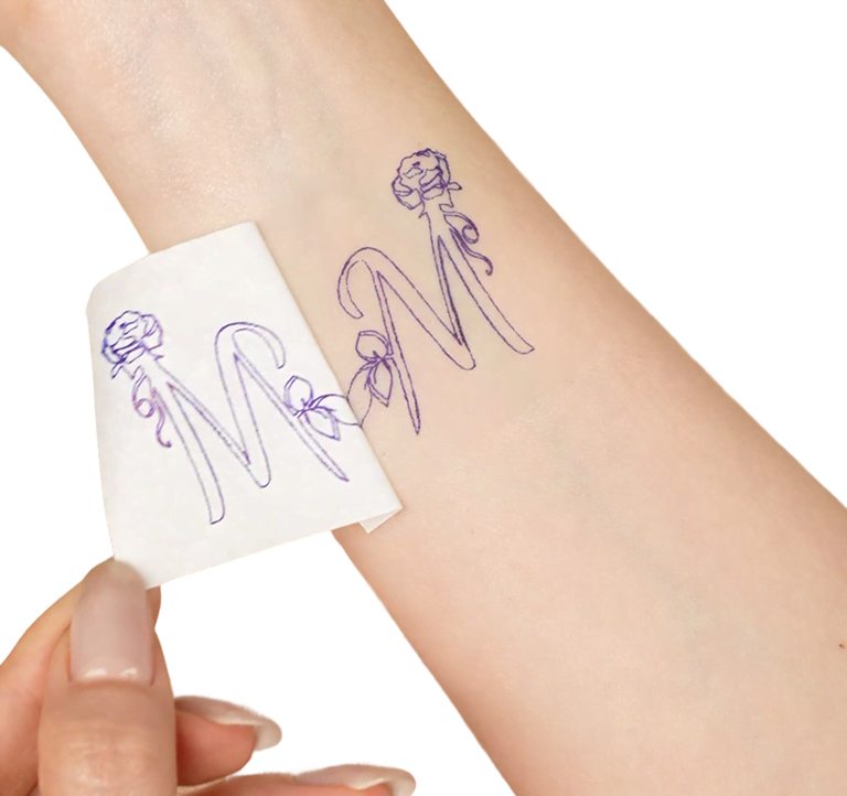 Mano femminile che stacca un pezzo di carta per tatuaggi con il disegno della lettera M e un piccolo fiore decorato.