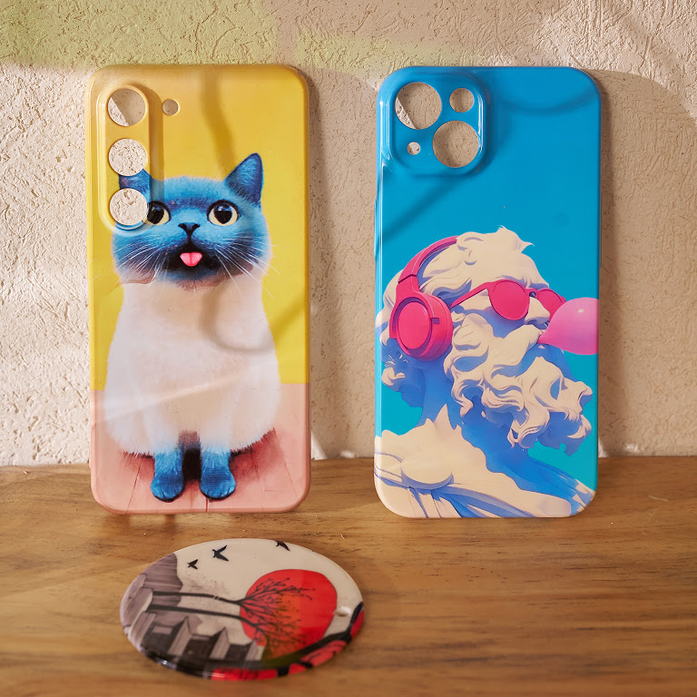Two phone cases on a shelf. One with an illustration of cat sticking out its tongue and the other of a statue with pink headphones and sunglasses blowing a pink bubble with bubblegum. In front of the cases is a button pin.