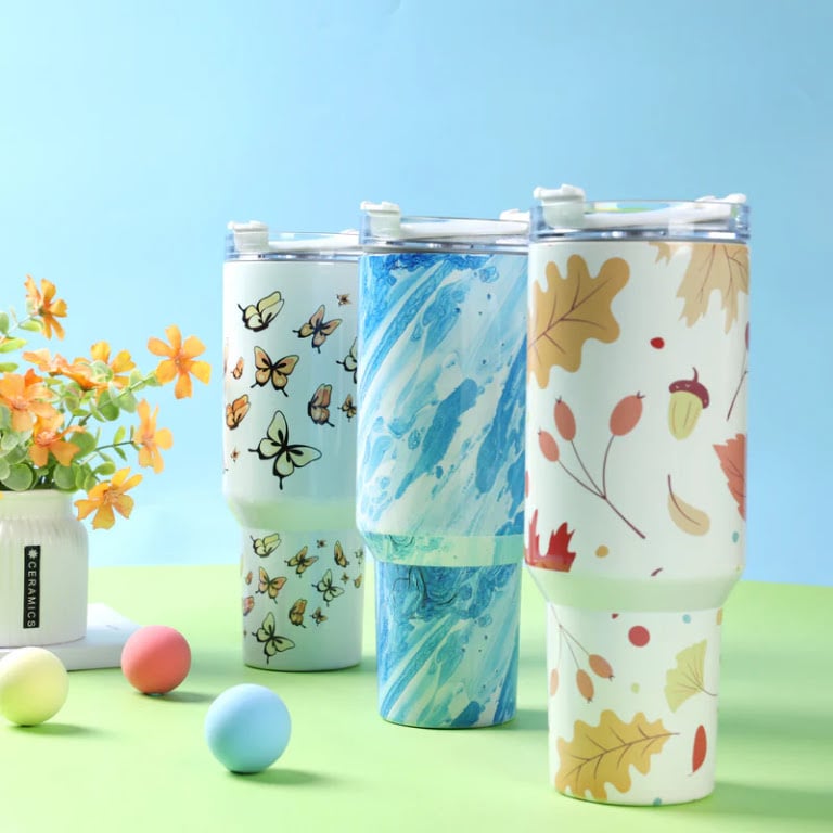 Three large stainless steel travel tumblers with handles showcasing custom sublimation wraps in butterfly, blue marble, and autumn leaf designs.