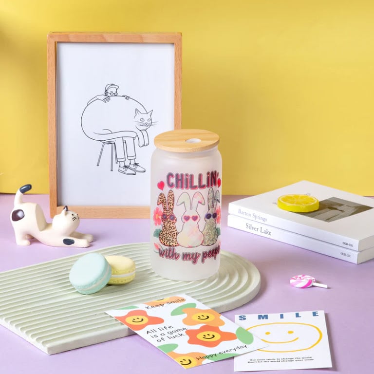 Frosted glass can tumbler with a bamboo lid featuring a custom sublimated bunny design, displayed with colourful pastel decor props.