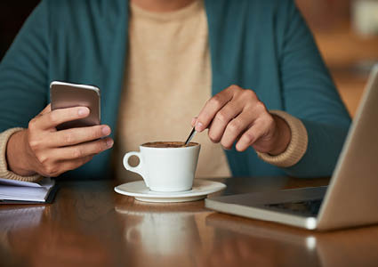 Iemand die aan de laptop zit met haar telefoon in de hand en een kop espresso