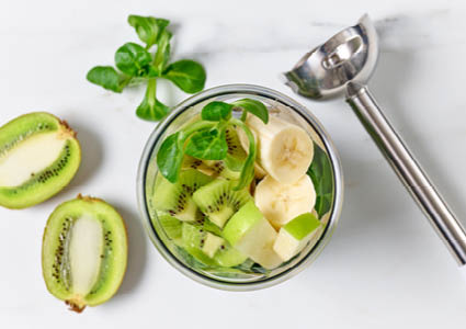 Een bovenaanzicht van een foodprocessor gevuld met bananen, kiwi's, appels en kruiden