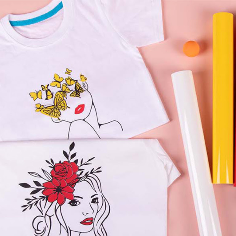 Top down view of two white T-shirts on a light pink background. The bottom shirt has a heat transfer print of a woman with red flowers in her hair. The top shirt shows a woman's face with the eyes obscured by yellow butterflies.