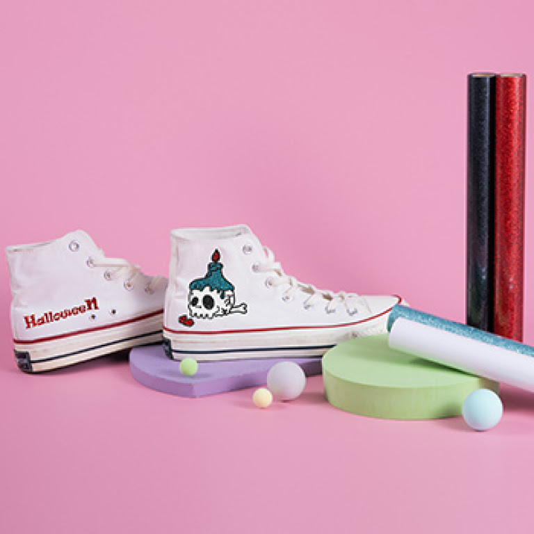 A pair of white canvas hi-top Converse shoes against a pink background. The shoes have a heat transfer print of a cartoon skull with a candle on top and the text Halloween. There are 4 rolls of LOKLiK glitter heat transfer vinyl.