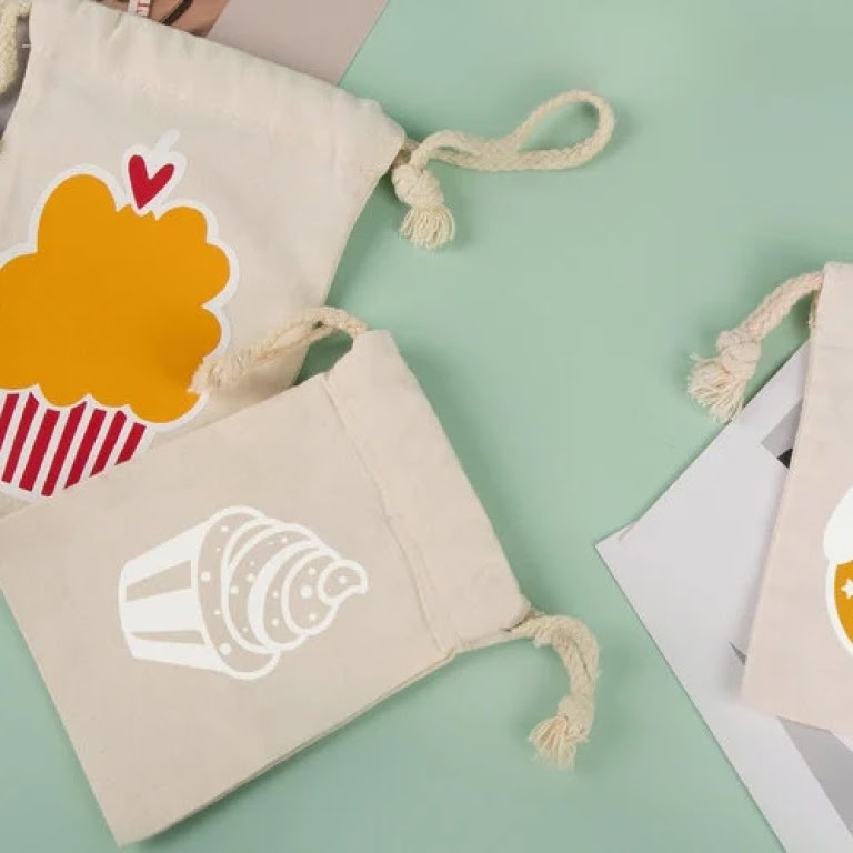 Top view of 3 small canvas drawstring bags on a light green background. The bags have cupcakes ironed on them.