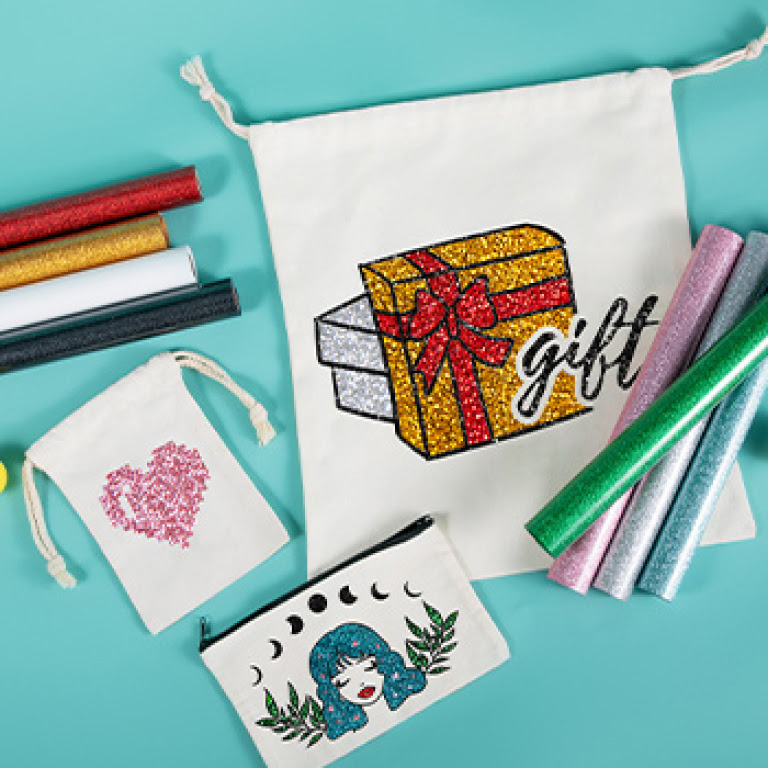 Top view of 3 canvas drawstring bags on an aqua background. They have a design of a heart, gift box and woman's face ironed on them in glitter heat transfer vinyl.