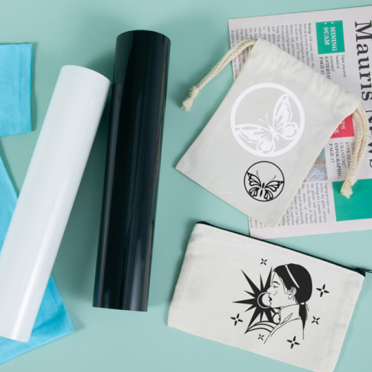 Tio down view of 2 white drawstring bags. One has a design of 2 butterlfies in white and black and the other a person seen from the side in black vinyl. There are 2 rolls of heat transfer vinyl in white and black.