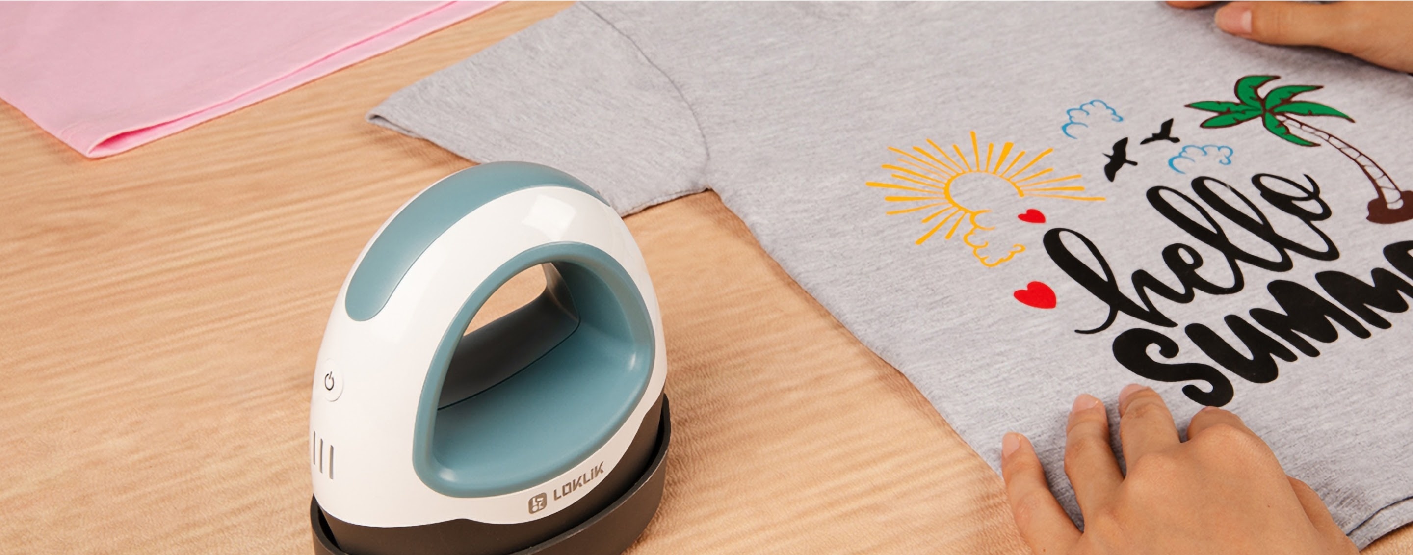 Table with a LOKLiK Mini Heat Press in Denim blue in its safety base. Next to it is a heather grey shirt with a heat transfer desing saying 'hello summer' in black, a yellow sun and a palm tree in green and borwn.