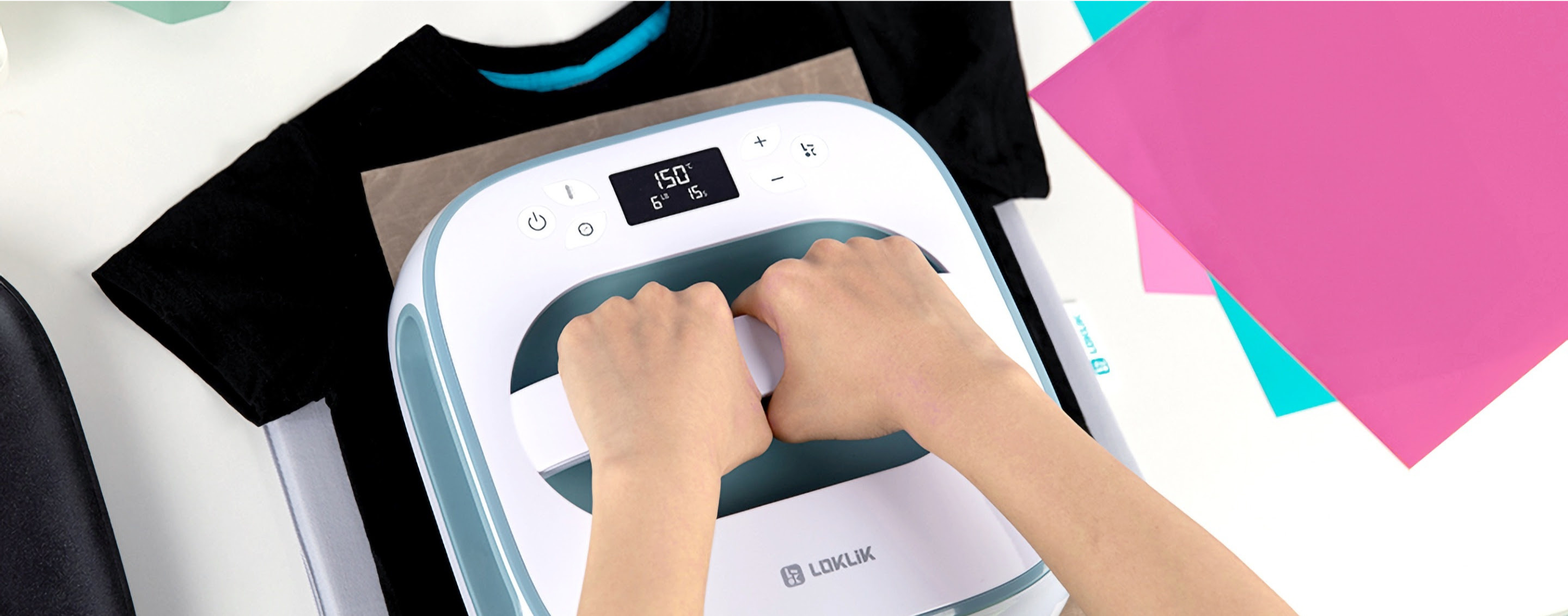 Two hands holding the top handle of a LOKLiK Easy Heat Press in Denim Blue. The Heat Press is ironing a design on a black shirt. On the right there are 3 sheets of heat transfer vinyl.