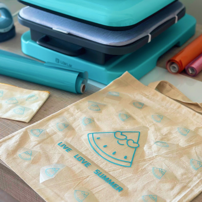 White tote bag with a cute watermelon design cut out of Aqua Blue Heat Transfer Vinyl and the text 'Live Love Summer'. In the background is a LOKLiK Auto Heat press and rolls of heat transfer vinyl.