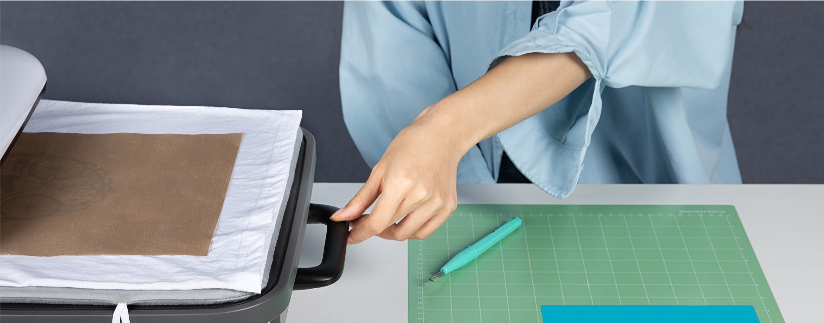 Woman in a light blue shirt reaching out and pulling out the drawer from a LOKLiK Auto Heat Press. There is a white shirt with a teflon sheet on top in the press. A green cutting mat and weeding tool are on the table.