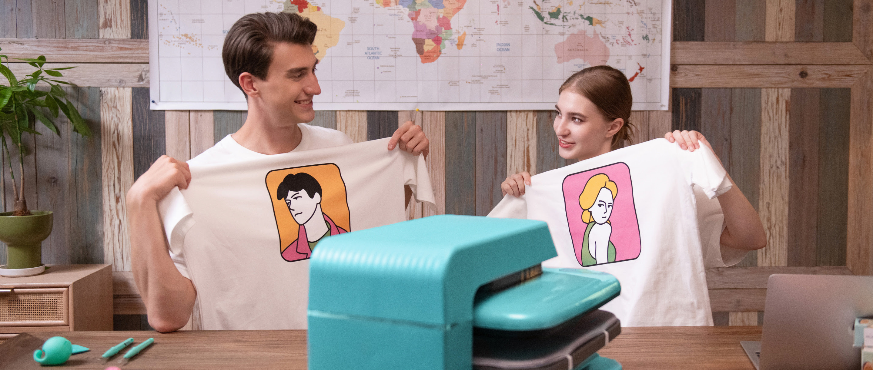 A man and a woman sitting behind a table looking at each other and each holding a white shirt with a heat transfer print of a lineart face. On the table is an Auto Heat Press in LOKLiK Blue. ON the wall is a poster with a map of the world.