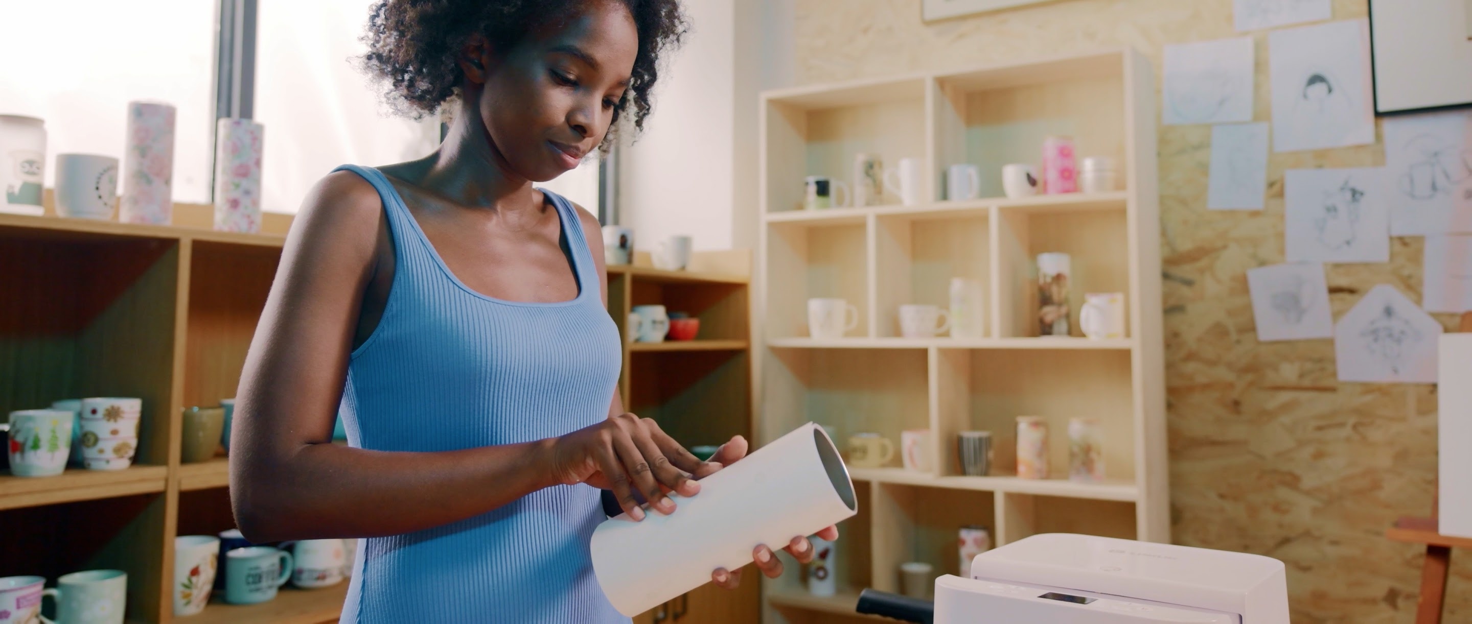 Woman of colour in a blue dress in front of shelves filled with mugs. She is wrapping a tumbler with sublimation paper.