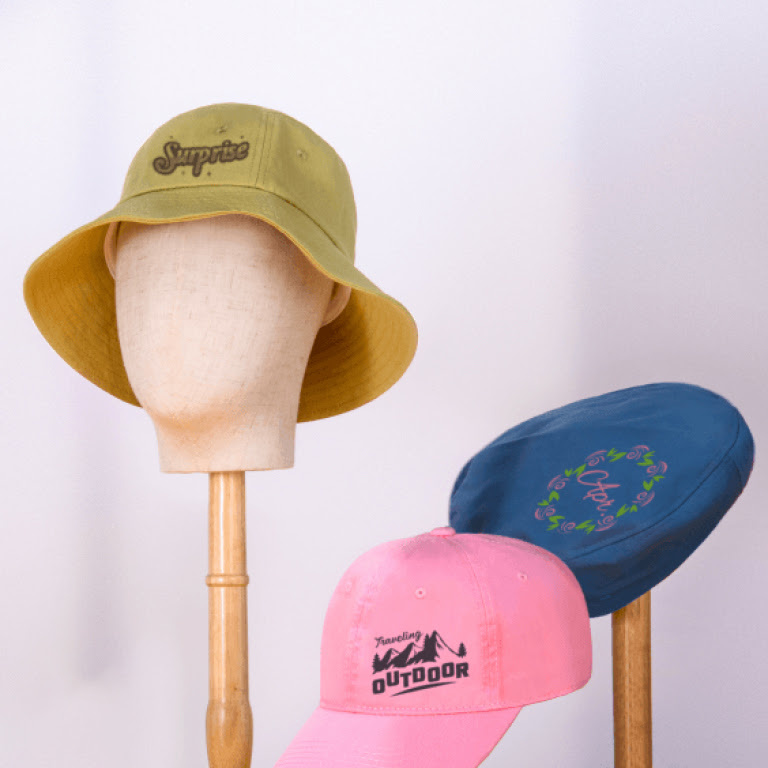Yellow bucket hat with an iron-on design of the words 'Surpirse', a blue flat cap with a wreath of flowers and a pink cap with moutains in black vinyl and the words 'outdoor'.