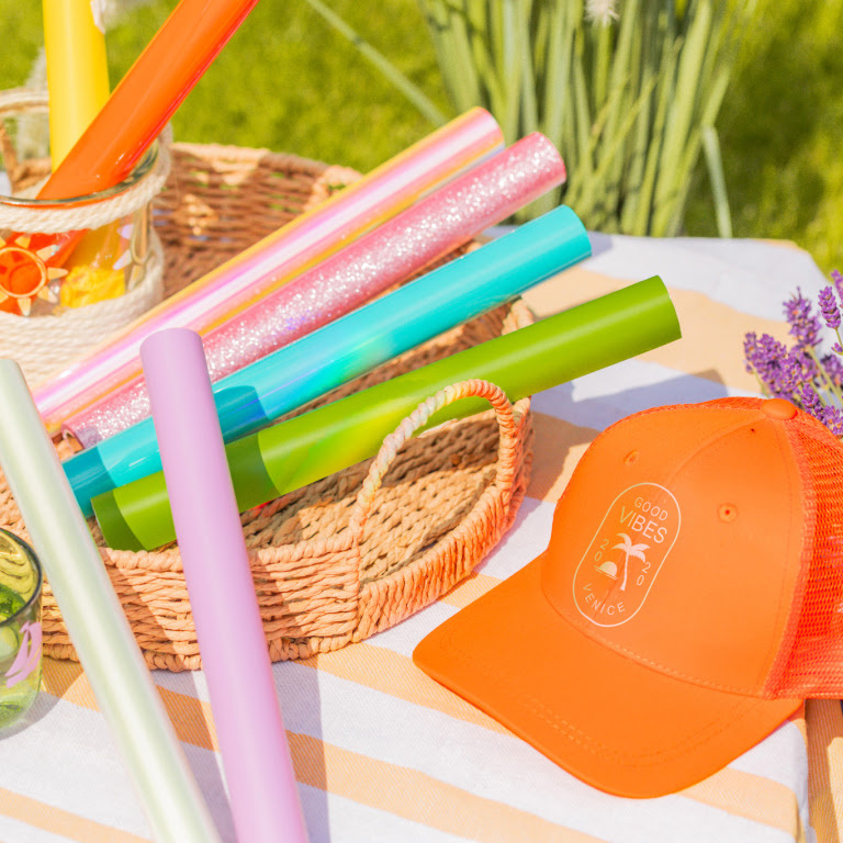 Orange cap with 'Good vibes' and palm tree ironed on. Several rolls of heat transfer vinyl are next to it.