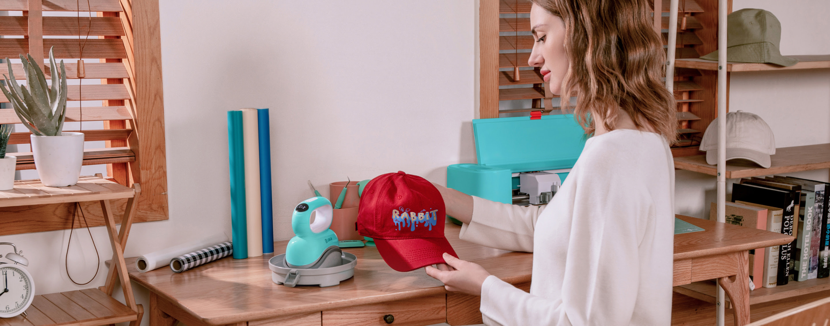 Woman at a desk holding a red cap with a vinyl design reading rabbit. On the table is a LOKLiK crafter, a LOKLiK Impress™ Hat and several rolls of heat transfer vinyl.