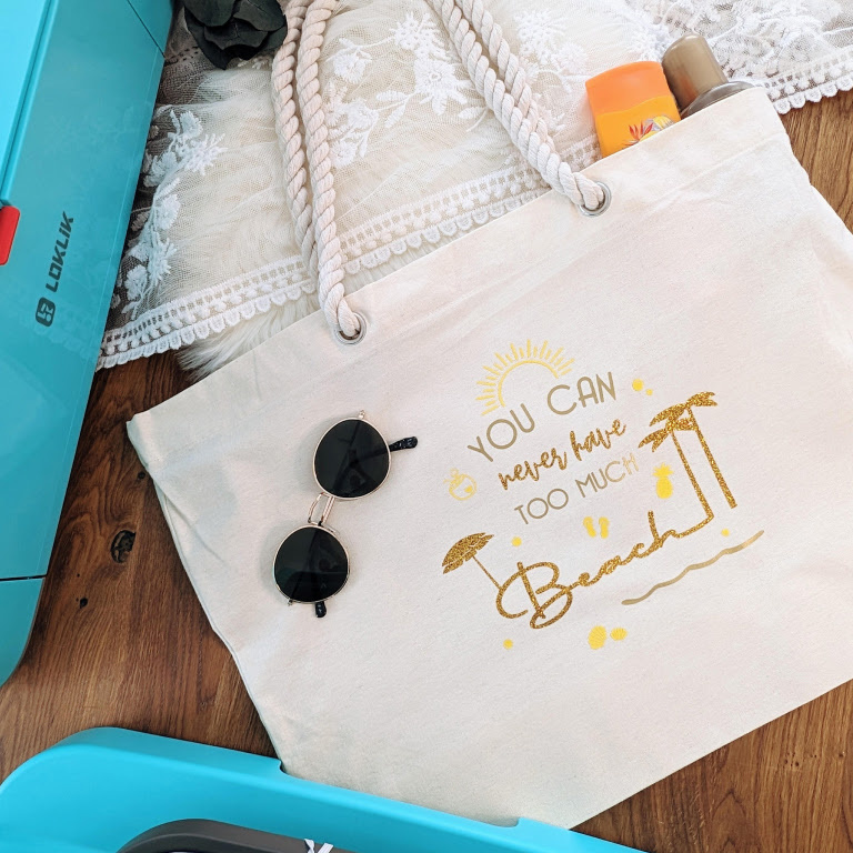 Top view of a white canvas bag with a desing in heat transfer vinyl with the text 'You can never have too much beach' and two palm trees and a parasol.