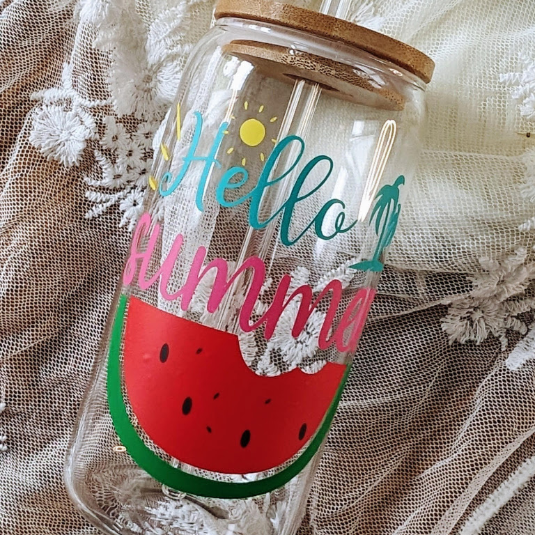 Glass jar lying on a table with sheer fabric. The jar has a design in permanent adhesive vinyl of a slice of watermelon and the text 'Hello Summer'