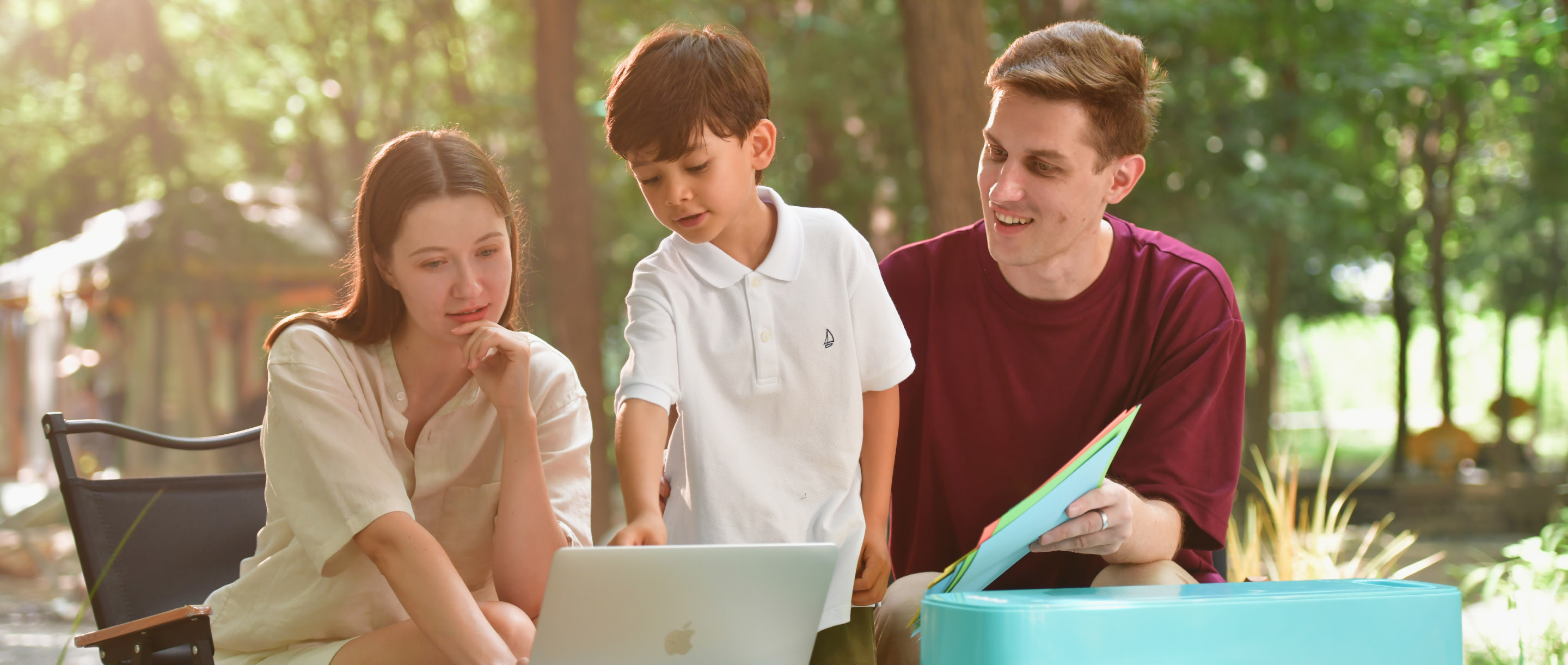 A family of three sitting at a laptop in a garden setting. On the table is a LOKLiK Crafter in LOKLiK Blue. The father is holding an array of colourful cardstock. The young child is pointing at the laptop.