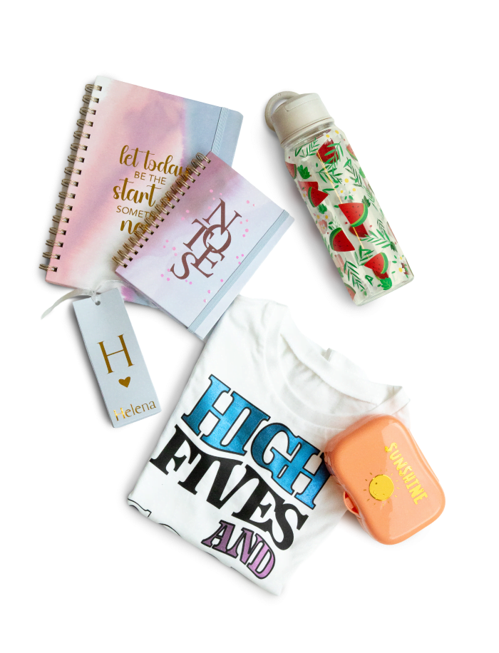 Top view of two notebooks with permanent adhesive letters. A plastic water bottle with a pattern of waternmelon and leaves made from permanent adhesive vinyl, a folded shirt with 'High fives and good vibes' in heat transfer  vinyl, a cardboard package with Helena in gold vinyl and a small salmon coloured plastic container with 'Sunshine' and a small sun in gold and yellow vinyl.