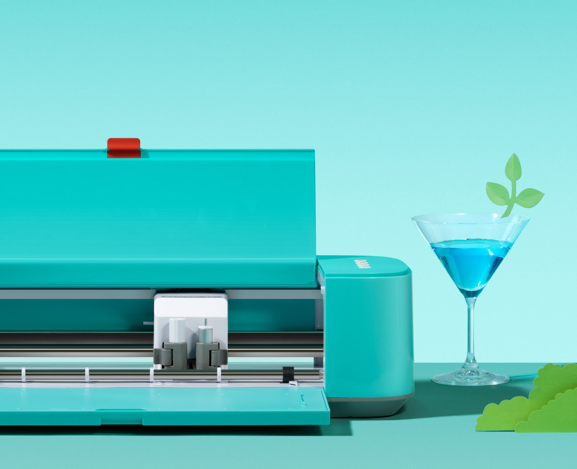 A LOKLiK Crafter in LOKLiK Blue in front of a light blue background on a teal table. The Crafter is open. On the right is a cocktail glass filled with a blue transparent liquid. It has a cut out shape of a small branch with 3 leaves in green cardstock stuck to the rim.