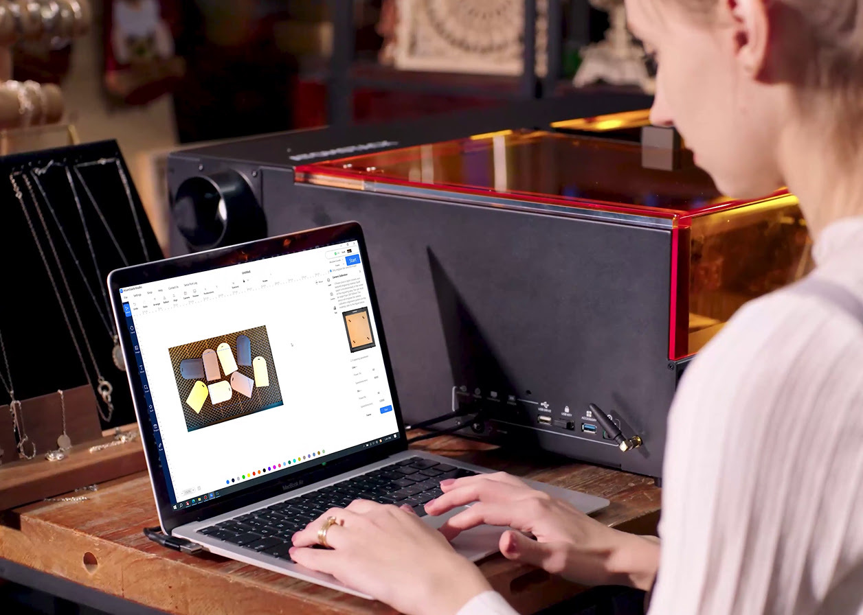 Woman working at a laptop showing the camera preview of the AtomStack Kraft in AtomStack Studio. Eight leather tags are scattered on the honeycomb.