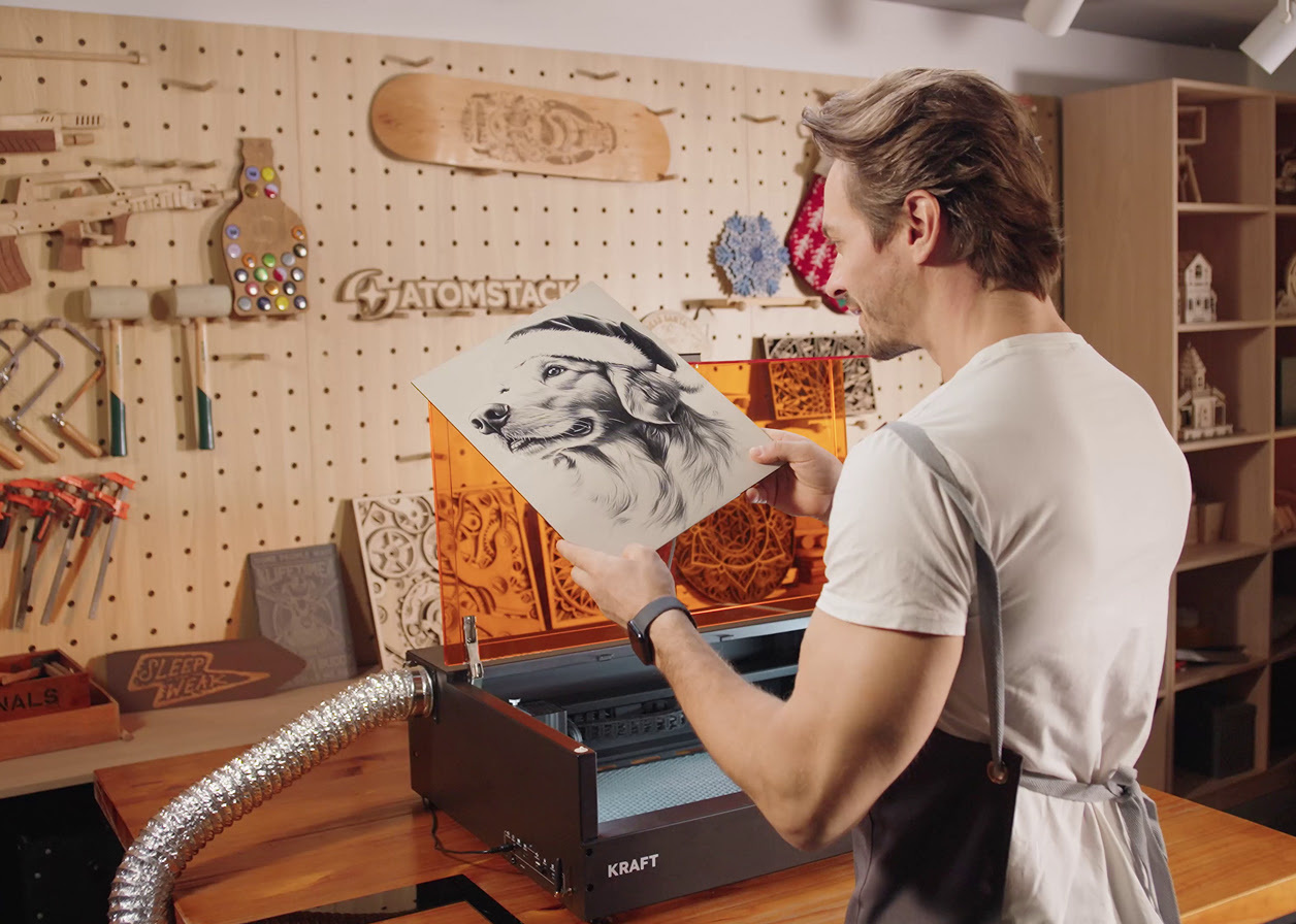 Man in a workshop holding a piece of wood engraved with a picture of a dog in a santa hat. In the background is an AtomStack Kraft with the lid open on a table.