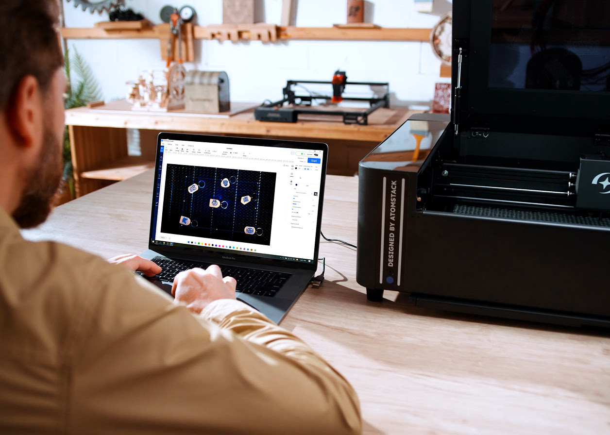 Looking over the shoulder of a man working on a laptop with AtomStack Studio open. The camera preview is showing 6 items on the honeycomb.