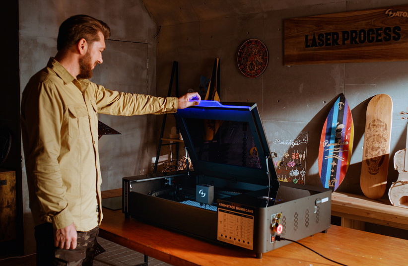 Man in a workshop opening an AtomStack Kraft on a wooden table. The blue led light of the Hurricane is on.