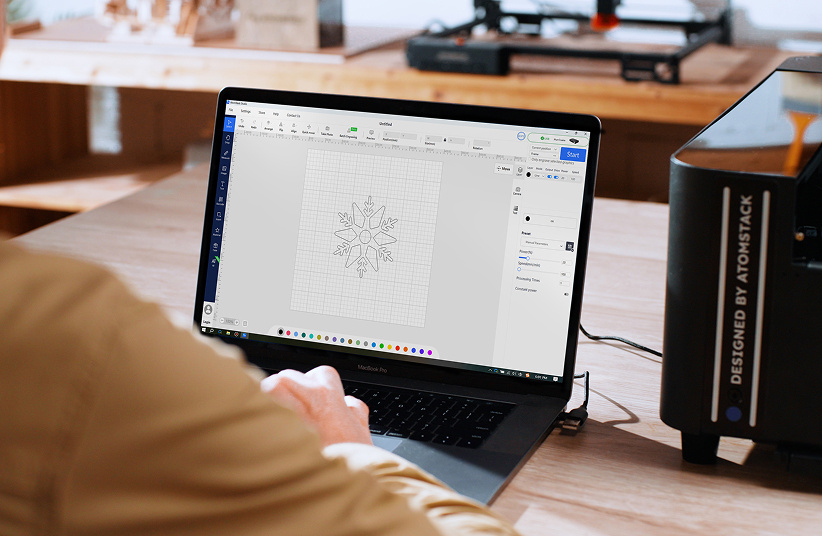 Looking over the shoulder of a man working on a laptop connected to an AtomStack Hurricane. AtomStack Studio is open showing a design of a snowflake.