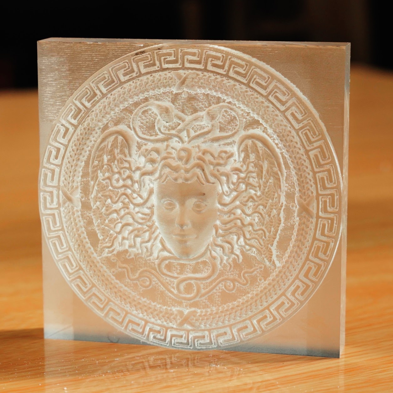 Square piece of acrylic standing upright on a wooden table. It has a milled circle with a pattern and the head of Medusa at the centre.