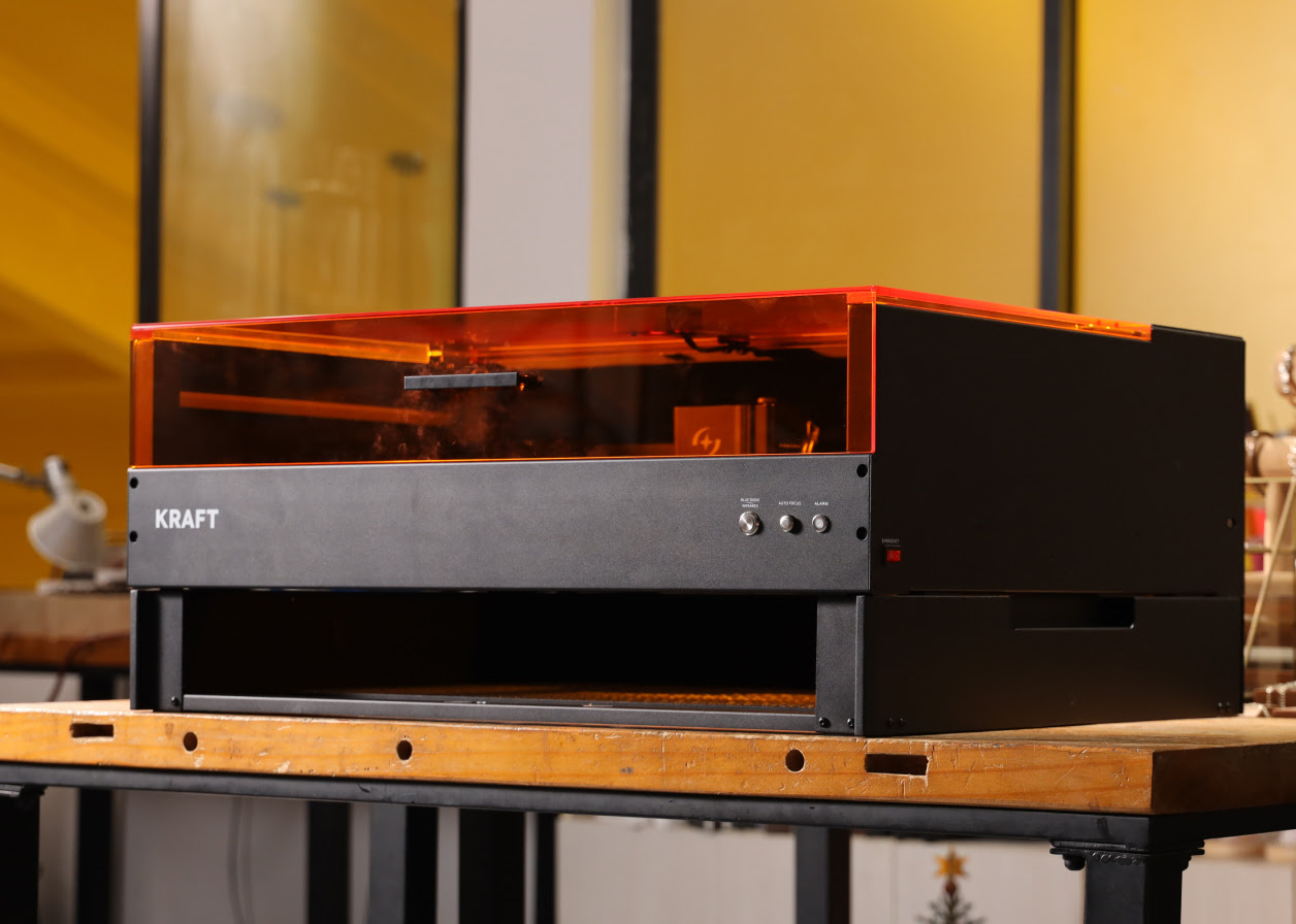 AtomStack Kraft with the riser base installed. The laser cutter is standing on a wooden table against a yellow background.