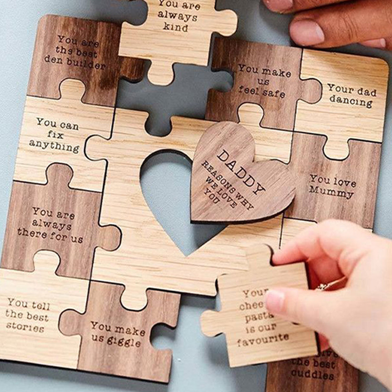 A laser-engraved wooden puzzle for Fathers Day featuring a central heart piece reading "Daddy reasons why we love you" surrounded by interlocking pieces with sweet messages being assembled by hands.