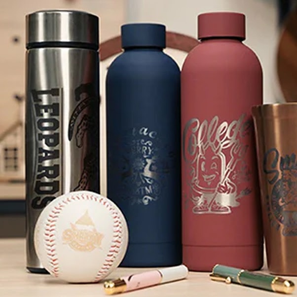 Custom laser-engraved stainless steel, dark blue, and matte red water bottles standing next to a laser-etched baseball and professional fountain pens on a desk.