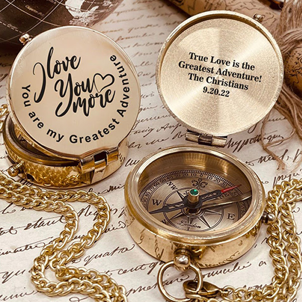 A laser-engraved pocket compass with an open lid, showing romantic custom inscriptions on both the outer casing and inner lid, resting on vintage cursive script paper with a gold chain.