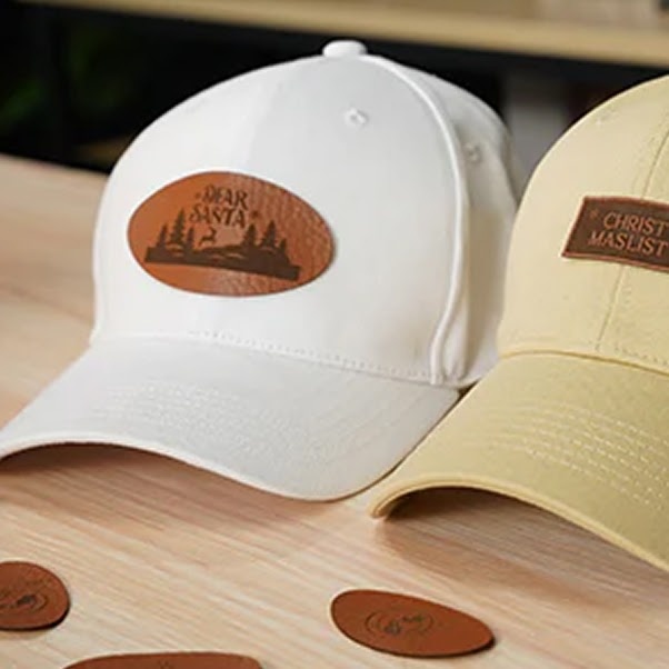 Custom baseball caps in white and beige featuring laser-engraved brown leather patches, with one oval patch reading Dear Santa alongside a pine tree silhouette and a rectangular patch reading Christmas List.
